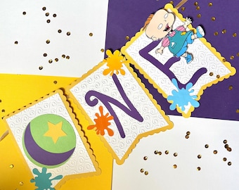 May include: A colorful banner with the word "ONE" and a cartoon character. The banner features a yellow border, white background with a swirl pattern, and purple letters. Decorative paint splatters and a cartoon character are also included.