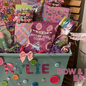 May include: A gift basket filled with treats and accessories. The basket is decorated with buttons and a bow, with the name "RYLIE" spelled out. Contents include lip balm, cookies, scrunchies, and a notebook. The text "BOW & BUTTONS BASKET!" is also visible.