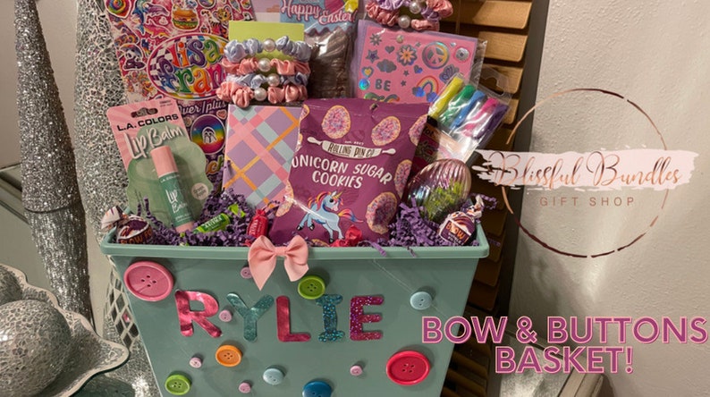 May include: A gift basket filled with treats and accessories. The basket is decorated with buttons and a bow, with the name "RYLIE" spelled out. Contents include lip balm, cookies, scrunchies, and a notebook. The text "BOW & BUTTONS BASKET!" is also visible.