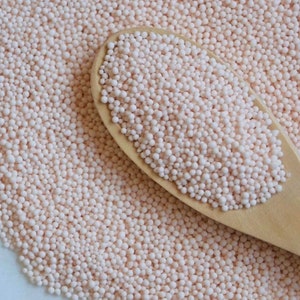 May include: A close-up of a wooden spoon filled with small, pink tapioca pearls. The pearls are scattered across a white surface.