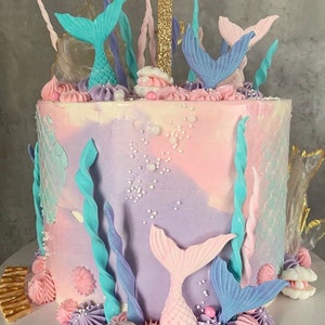 以下が含まれることがあります： パステルピンクとパープルのケーキは、人魚の尻尾、貝殻、海藻で飾られています。ケーキの上には、金色のグリッターでできた数字の1が飾られています。