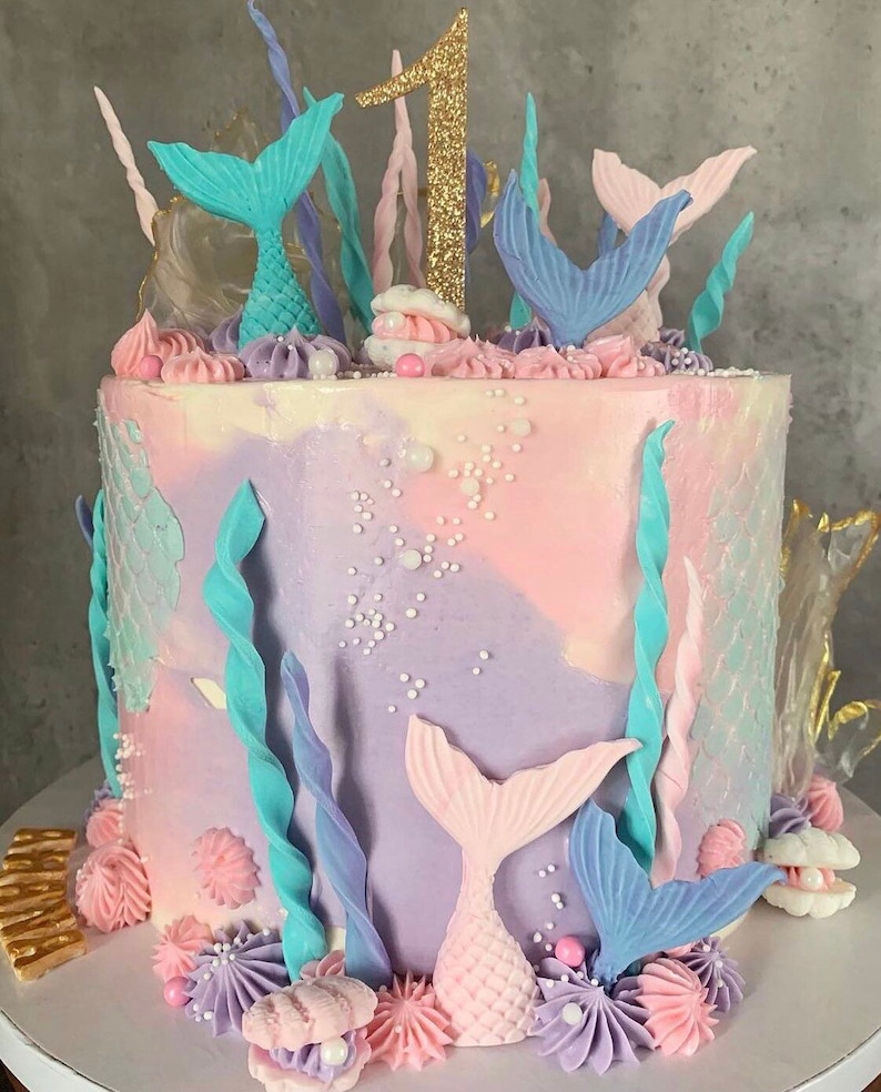 以下が含まれることがあります： パステルピンクとパープルのケーキは、人魚の尻尾、貝殻、海藻で飾られています。ケーキの上には、金色のグリッターでできた数字の1が飾られています。