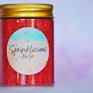 May include: A glass jar with a gold lid filled with red sprinkles. The jar has a white round label with the text "Sprinklicious by Lis" in black.