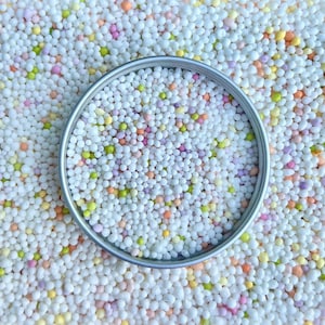 May include: A close-up of a white, round container filled with colourful sprinkles. The sprinkles are mostly white with small amounts of pink, orange, and green.