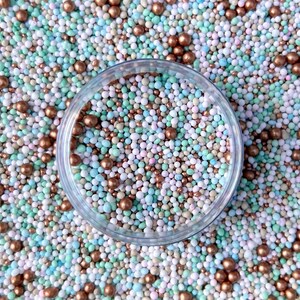 May include: A close-up of a clear glass bowl filled with colorful sprinkles. The sprinkles are a mix of small, round beads in shades of blue, green, pink, white, and gold. The bowl is sitting on a bed of the same sprinkles.