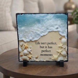 May include: A decorative square plaque featuring a beach scene with a blue ocean and white waves. The plaque includes seashells and starfish, with the text "Life isn't perfect, but it has perfect moments."