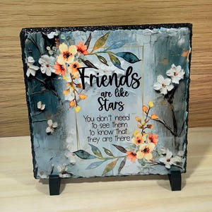 May include: Square decorative plaque with the text "Friends are like Stars" and a floral design in orange, white, and blue. The plaque is supported by a black stand and has a textured, stone-like appearance.