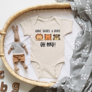 May include: A beige baby onesie with the text "Lions, Tigers, & Bears, OH MY!" and cartoon illustrations of a lion, tiger, and bear. The onesie is in a woven basket with a stuffed bunny and a gray blanket.
