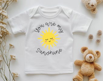 Może przedstawiać: Białe body niemowlęce z napisem "You are my sunshine" i żółtym uśmiechniętym słońcem. Na zdjęciu znajduje się również pluszowy miś i dekoracyjne kwiaty.