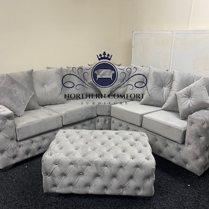 May include: A gray sectional sofa with a matching ottoman. The sofa has a button-tufted design on the arms and base, with several plush cushions. The ottoman is also button-tufted. The logo "NORTHERN COMFORT FURNITURE" is visible.