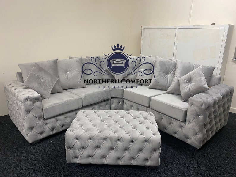 May include: A gray sectional sofa with a matching ottoman. The sofa has a button-tufted design on the arms and base, with several plush cushions. The ottoman is also button-tufted. The logo "NORTHERN COMFORT FURNITURE" is visible.