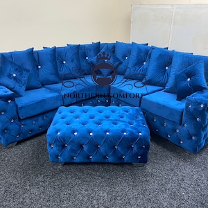 May include: A luxurious, blue velvet sectional sofa with a matching ottoman. The sofa and ottoman feature button tufting and crystal accents. The sofa includes several matching throw pillows. The logo "NORTHERN COMFORT" is visible.