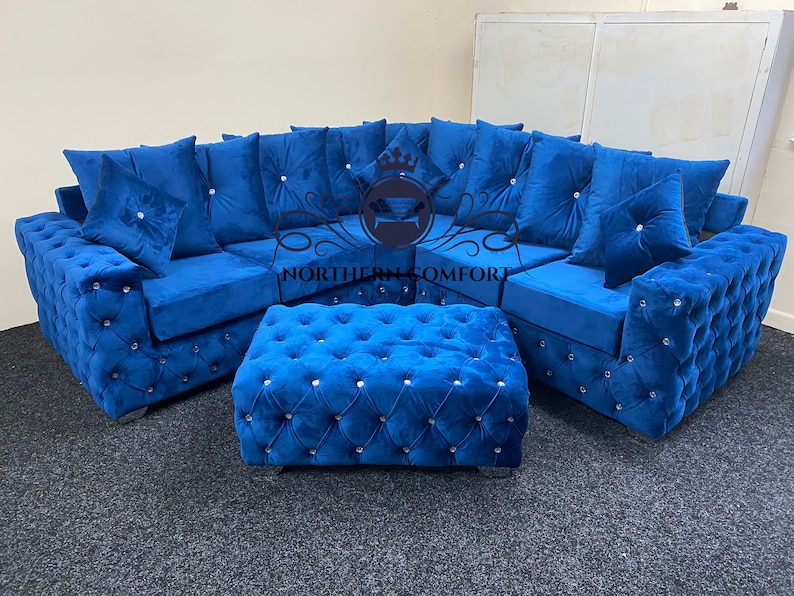 May include: A luxurious, blue velvet sectional sofa with a matching ottoman. The sofa and ottoman feature button tufting and crystal accents. The sofa includes several matching throw pillows. The logo "NORTHERN COMFORT" is visible.