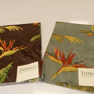 May include: Two fabric swatches with a floral pattern featuring bird of paradise flowers. One swatch is brown, the other is green. Both have yellow, orange, and green floral designs. Each swatch has a white label with the text "THIBAUT Wallpaper & Fabrics Since 1886."