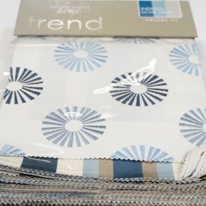 Puede incluir: Una pila de muestras de tela con una variedad de patrones, incluyendo una tela blanca con diseños de rayos de sol azules y grises, y una tela a rayas con rayas azules, grises y beige. Las muestras de tela están etiquetadas como "Trend Home Fabrics".