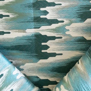 May include: Close-up of a fabric with a repeating geometric pattern in shades of teal, white, and gray. The design features a series of horizontal, abstract shapes with a textured appearance. The fabric is folded, revealing the pattern on both sides.
