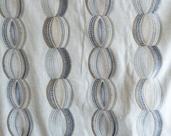 May include: Close-up of a cream-colored fabric with an embroidered pattern. The pattern features vertical rows of connected oval shapes in shades of blue and beige, creating a textured design. The fabric appears to be linen or a similar material.