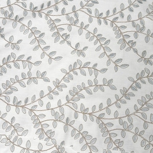 May include: A white fabric featuring an embroidered pattern of winding vines with light blue leaves and tan stems. The repeating botanical design covers the entire surface, showcasing intricate stitching that creates a textured appearance.