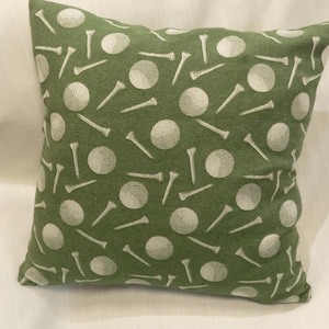 May include: Square green decorative pillow with a repeating pattern of white golf balls and tees. The pillow is made of a soft fabric and is likely used for home decor. The pillow is square and measures approximately 16 inches by 16 inches.