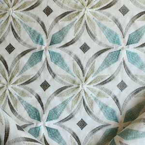 May include: White fabric with an embroidered geometric pattern in shades of teal, brown, and beige. The pattern features repeating diamond shapes and stylized flower motifs.