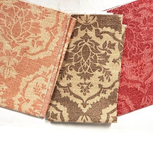 May include: Three rectangular fabric swatches featuring a repeating damask pattern. The left swatch is peach and cream, the center swatch is brown and beige, and the right swatch is a solid deep red with a tonal damask print texture.