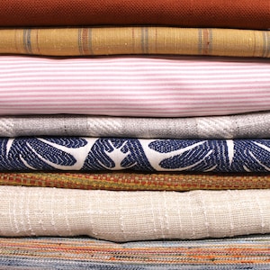 May include: A stack of fabric swatches in various colours and patterns, including a rust-coloured fabric, a striped fabric, a pink and white striped fabric, a grey fabric, a blue floral fabric, a brown fabric, and a beige fabric.