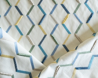May include: Close-up of a white fabric with a geometric pattern. The pattern features zig-zag shapes embroidered in shades of blue, green, yellow, and silver. The fabric is folded, revealing the pattern's repeating design, suitable for home decor.