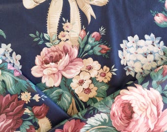 May include: Close-up of a navy blue floral fabric with a repeating pattern of pink roses, white hydrangeas, and cream-colored bows. The fabric also features smaller white and cream-colored flowers and green leaves. The overall design is a classic, vintage style.