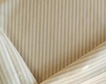 Puede incluir: Primer plano de una tela con un patrón de rayas verticales. Las rayas alternan entre beige claro y un gris apagado. La tela está doblada en las esquinas, mostrando la textura y la dirección del tejido.