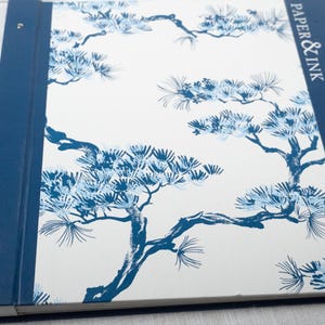May include: A rectangular scrapbook with a white cover featuring a blue pine tree branch pattern. The spine and side border are solid blue with the text PAPER & INK and Natural Silhouettes printed in white. A silver rope handle is attached.