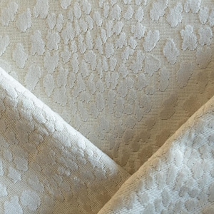 Puede incluir: Primer plano de una tela crema doblada con un patrón repetitivo de formas irregulares en relieve. La tela tiene un aspecto texturizado, con el patrón creando una superficie sutil y táctil. Los pliegues añaden profundidad al textil.