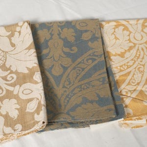 May include: Three folded fabric swatches featuring a repeating damask pattern. The left swatch is beige and cream, the center swatch is slate blue and beige, and the right swatch is mustard yellow and cream. Each piece has a textured weave.