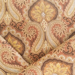 May include: Close-up of a folded fabric with a paisley pattern. The design features intricate floral and teardrop shapes in shades of brown, gold, and cream. The fabric has a textured appearance.