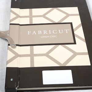 May include: A black and beige fabric sample binder featuring a geometric diamond pattern on the cover. The text FABRICUT LINEN CHIC is printed in white on a rectangular beige label. A small beige handle is attached to the side of the binder.