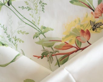 May include: Close-up of a white fabric with a floral pattern. The design features green leaves, yellow and orange flowers, and a black and white bee. The fabric is folded, revealing a plain white underside.