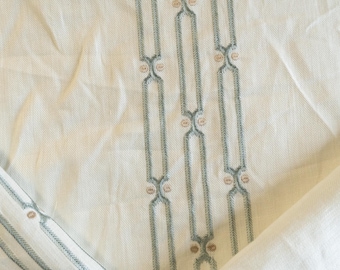 Puede incluir: Tela color crema con diseños bordados verticales en verde azulado y marrón claro. La tela tiene una textura sutil y está doblada, mostrando las costuras detalladas y las variaciones de color. El diseño es un patrón repetitivo.