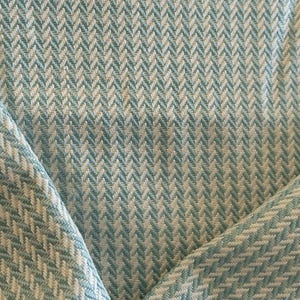 May include: Close-up of a woven fabric with a repeating herringbone pattern. The fabric features a combination of light blue and off-white threads, creating a textured surface. The fabric is folded, showing the pattern's direction.