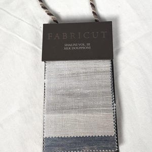 May include: A Fabricut Shalini Vol. III silk douppioni fabric sample swatch card featuring three horizontal rectangular sections of textured fabric in shades of light grey, beige, and dark blue, attached to a dark brown header card with a hanging cord.