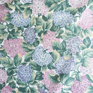 May include: A floral pattern featuring clusters of pink, purple, and blue hydrangea blooms with green leaves. The design is set against a white background, creating a dense, all-over botanical print. The pattern is suitable for fabric or wallpaper.
