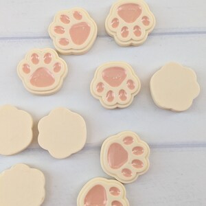 May include: Assortment of eight small, round, cream-colored resin paw print buttons with pink paw prints.