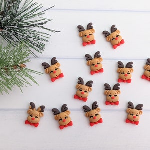 May include: Twelve brown reindeer head ornaments with red bows. The reindeer have brown antlers, brown eyes, and red noses. The ornaments are on a white wooden background.