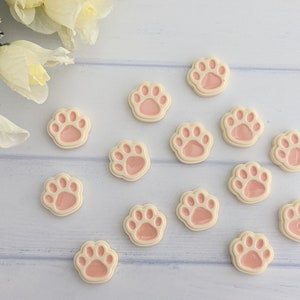 May include: A collection of 14 pink and white paw print shaped buttons. The buttons are arranged in a semi-circle on a white surface.