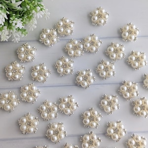 May include: A collection of 25 pearl and rhinestone embellishments in the shape of flowers. The embellishments are silver and white and have a delicate, feminine design.