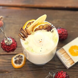 May include: A festive holiday scene featuring a clear glass filled with a creamy white beverage, garnished with dried citrus, cinnamon sticks, and a snowman. Red baubles and a matchbox with orange illustrations are scattered on a wooden surface.