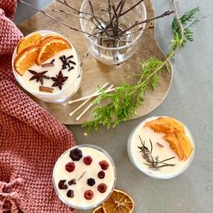 May include: Three glass candle jars with different scents and decorations. The candles are topped with dried orange slices, cinnamon sticks, star anise, and berries.