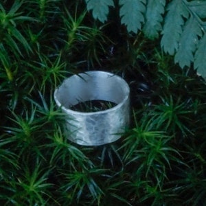 May include: A silver hammered ring with a wide band, resting on a bed of green moss.
