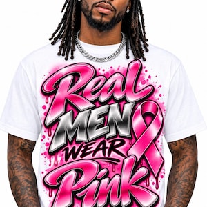 May include: A white short-sleeved t-shirt featuring airbrushed-style graffiti lettering in pink, black, and silver. The text reads Real MEN WEAR Pink, accompanied by a large pink awareness ribbon graphic with pink paint splatter accents.