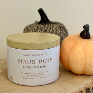 May include: A white cylindrical soy wax candle with a light wood grain lid. The label reads NIGHTROSE & CO, SOUS-BOIS DEEP WOODS, CHANDELLE DE SOYA SOY WAX CANDLE, 85 g, 15-20 h, and ST-JOSEPH-DU-LAC. Two decorative pumpkins sit behind the candle.