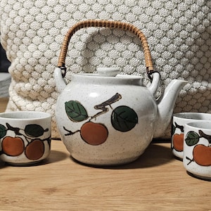 May include: A ceramic tea set consisting of a round teapot with a woven rattan handle and four matching handleless cups. The off-white speckled pottery features a hand-painted design of brown fruit and green leaves on each piece.
