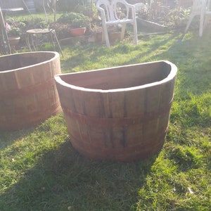 May include: Two large, weathered wooden planters with metal bands sit on a grassy lawn. One planter is a full circle, the other is a half-circle shape. The planters are brown and appear to be aged.